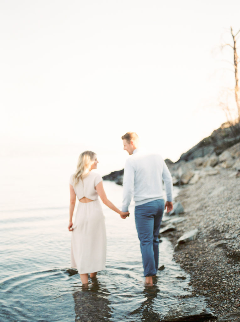an engaged couple hold hands and walk through Kelowna Lake 