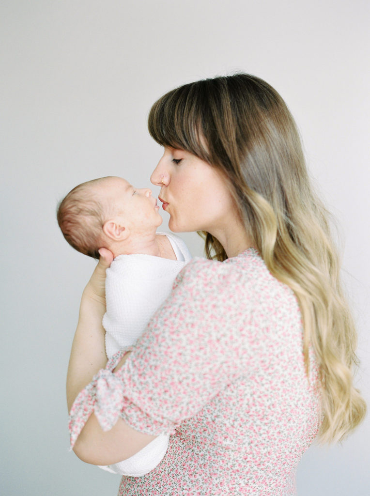 mom in pink dress kisses newborn baby boy 
