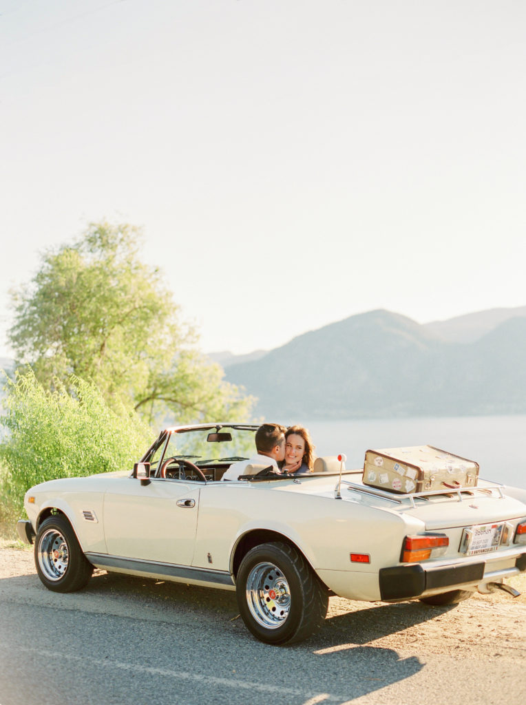 a man kisses a woman in a convertible parked on a hill 