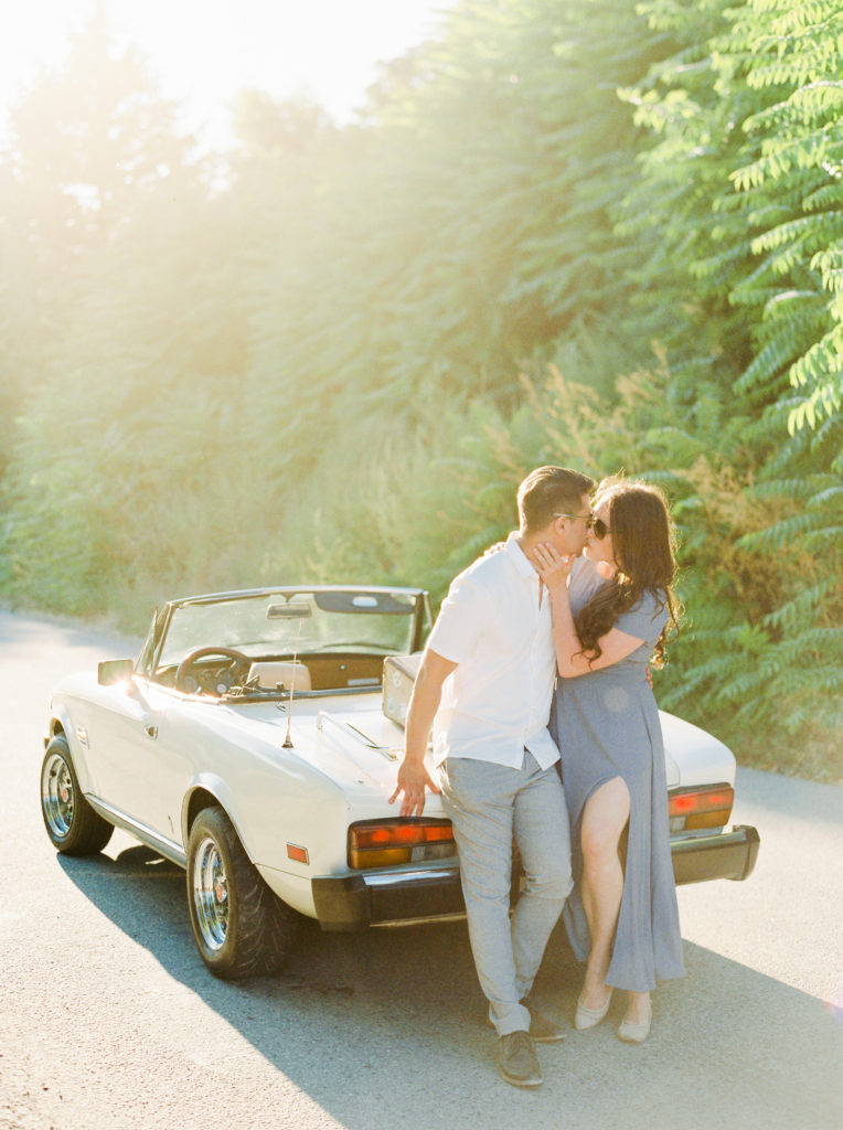 an engaged couple kiss while leaning on a classic Fiat convertible 
