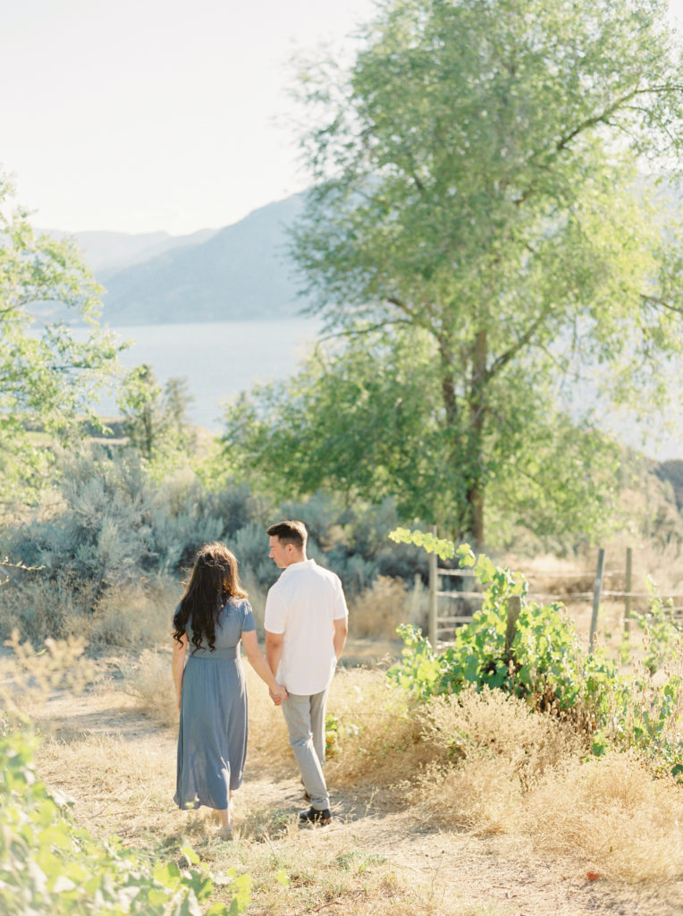 a man and woman hold hands and walk through a Naramata vineyard